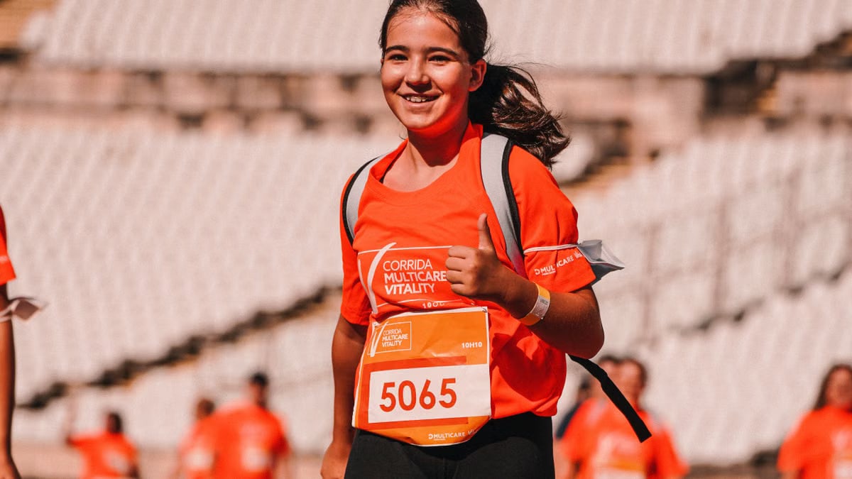 Smiling girl running in a marathon, giving a thumbs up, wearing a bright orange shirt.